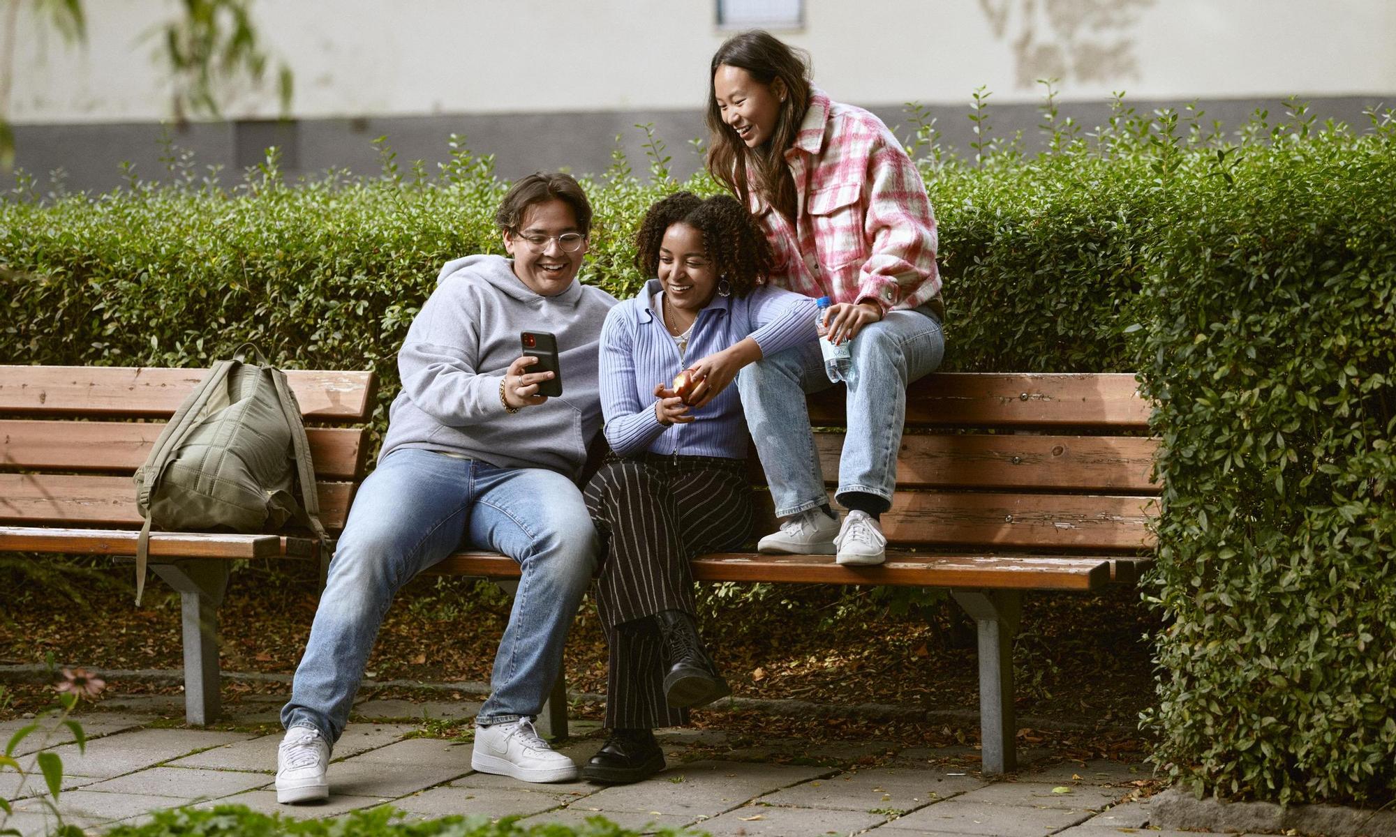 Three young persons on a bench laughing at a mobile