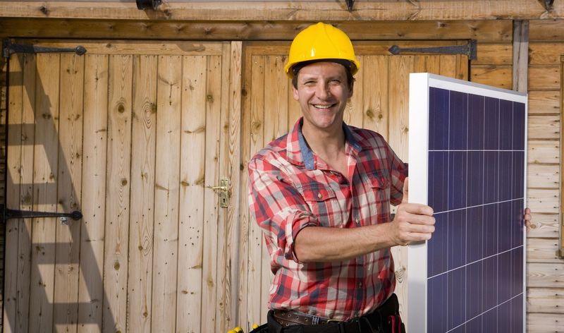 Smiling man holding solar panel in front of cabin